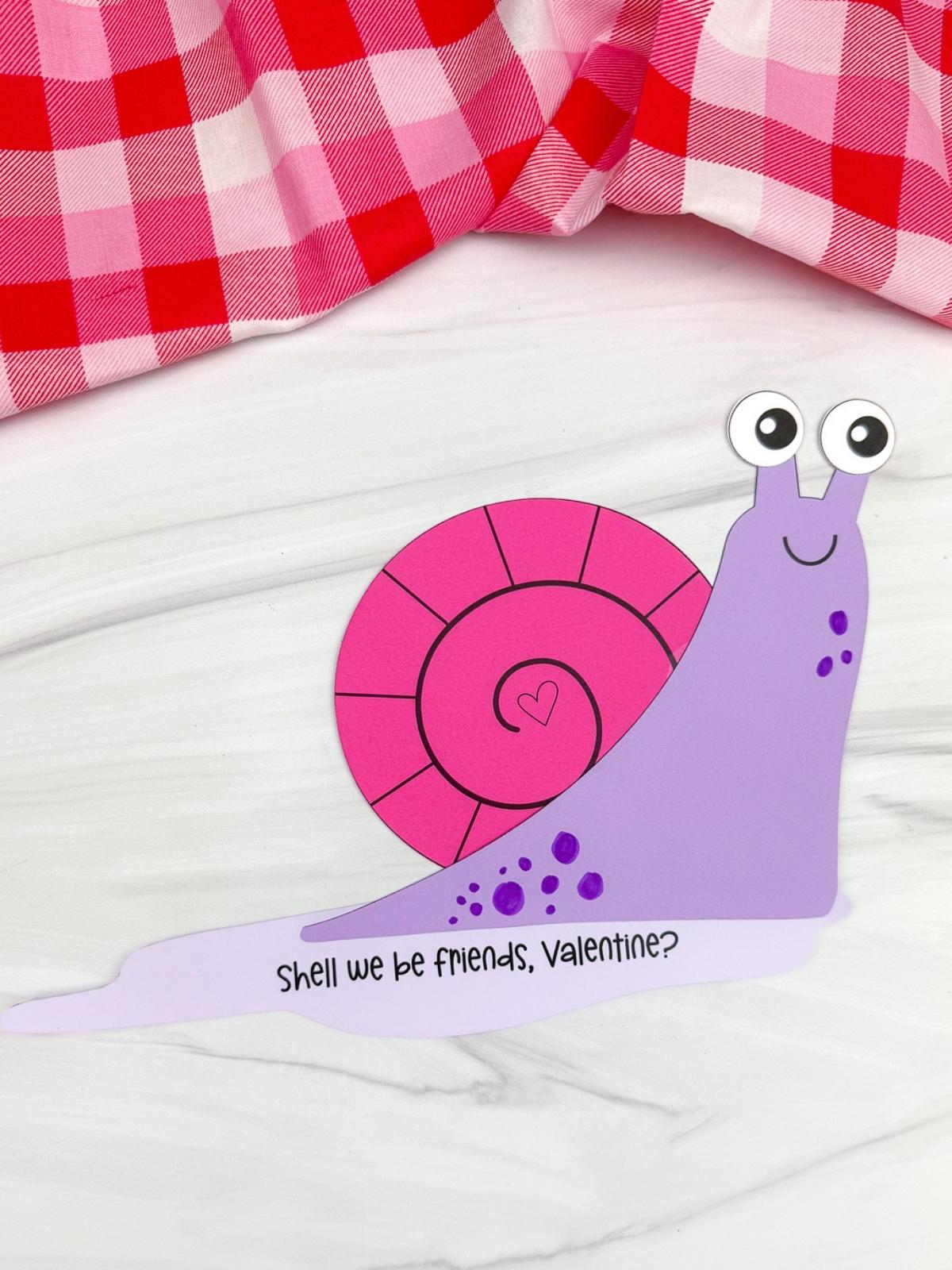 A completed Craft showing a purple paper Valentine snail with a pink heart shell googly eyes and the message Shell we be friends Valentine on a white background.