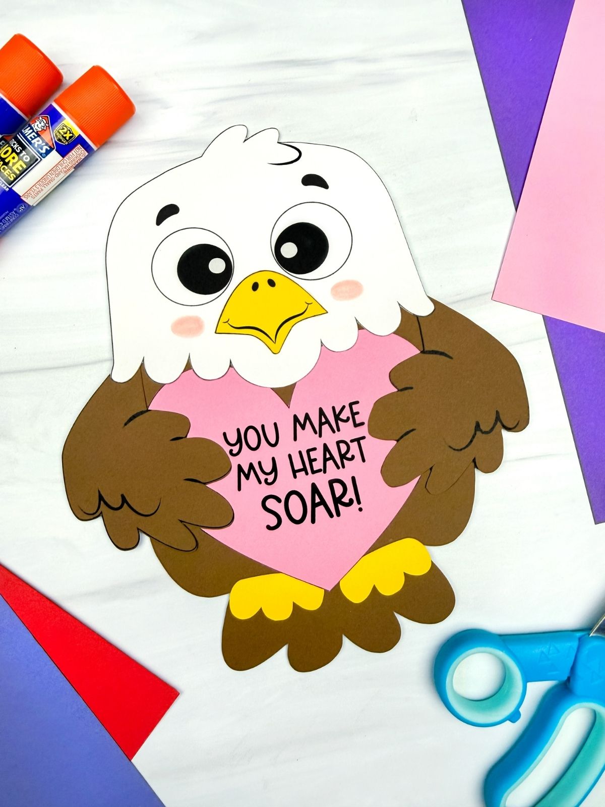 A completed craft of a cute Valentine eagle holding a pink heart with the message “You make my heart soar,” surrounded by colorful paper, glue sticks, and scissors.
