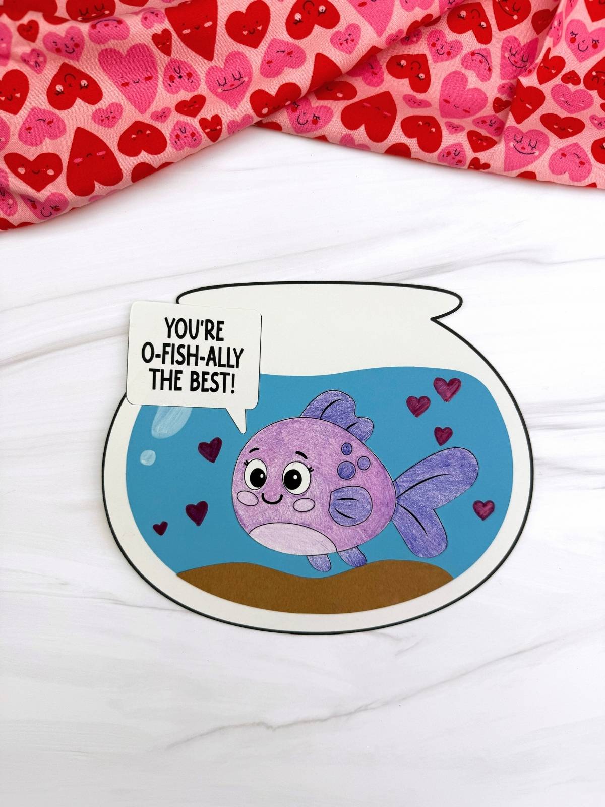 A completed craft of a Valentine fish bowl card with a purple fish, heart decorations, and a speech bubble saying you’re o-fish-ally the best.