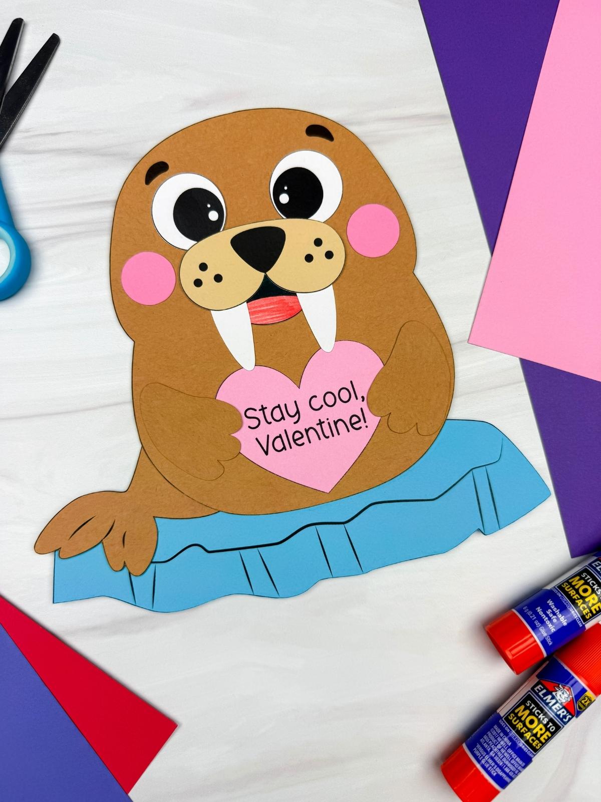 A completed craft showing a paper walrus Valentine holding a pink heart that reads Stay cool Valentine, sitting on a blue ice floe with scissors, glue sticks, and colorful cardstock for an easy kids Valentine craft.
