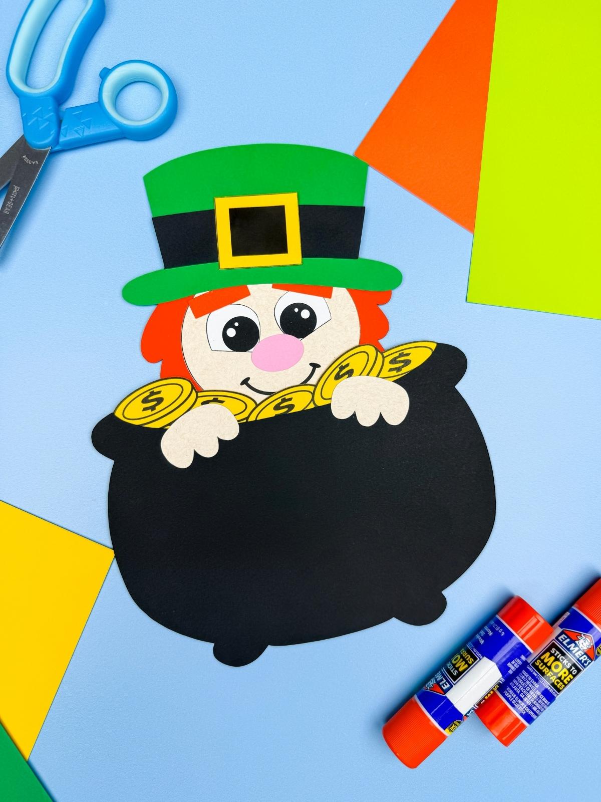 A completed Craft of Leprechaun in a Pot of Gold Craft with green hat and gold coins displayed with scissors, glue sticks, and colorful paper for a St. Patrick’s Day kids craft.