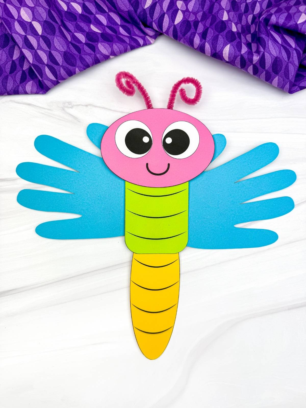A completed craft showing a colorful dragonfly handprint paper craft with blue wings and pink antennae, a fun and easy spring insect craft for preschool and kindergarten kids.
