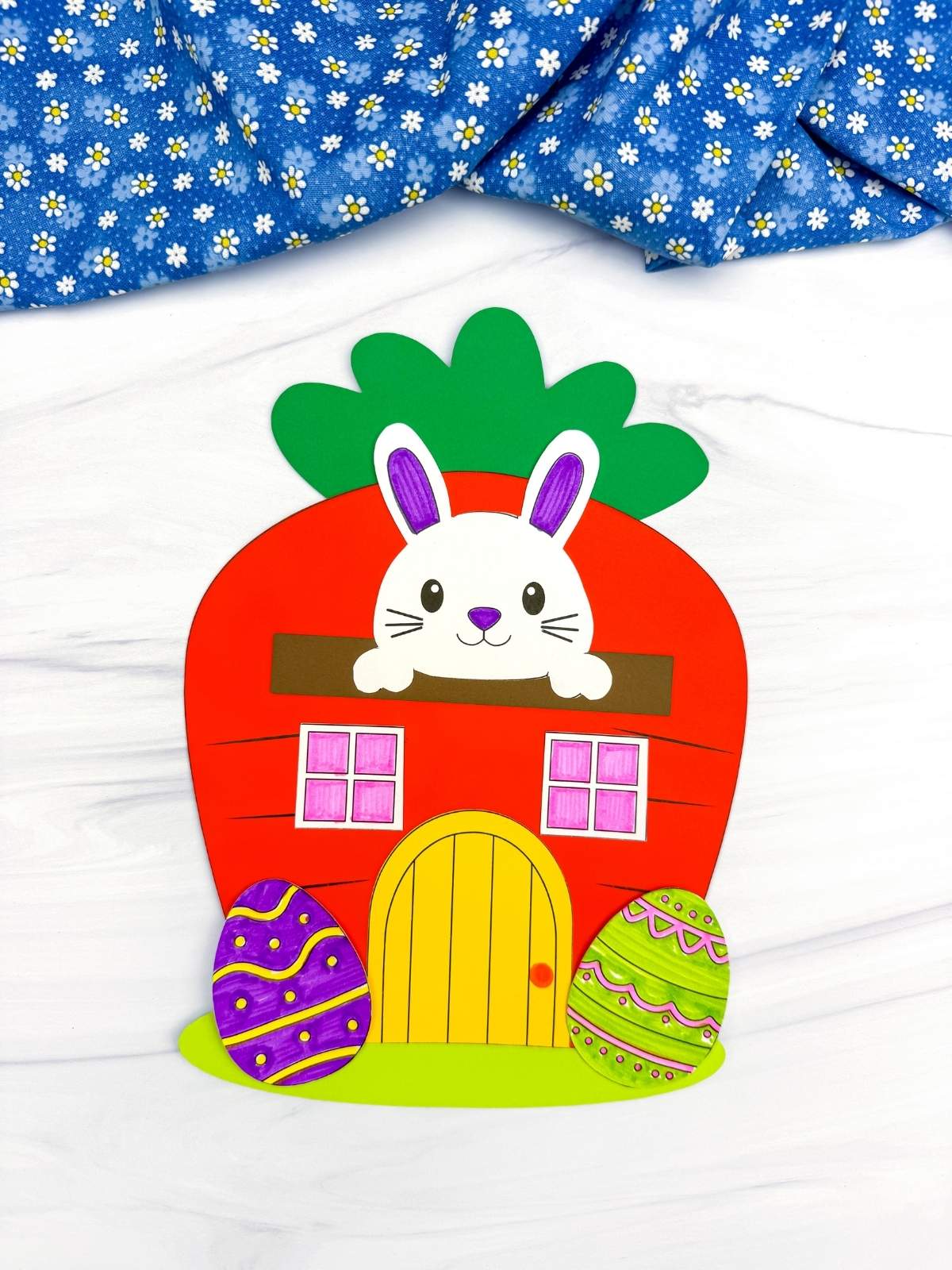 A completed Craft Easter Bunny House made from a carrot shape with a bunny peeking out, colorful decorated Easter eggs, and bright paper details for a fun kids Easter craft.