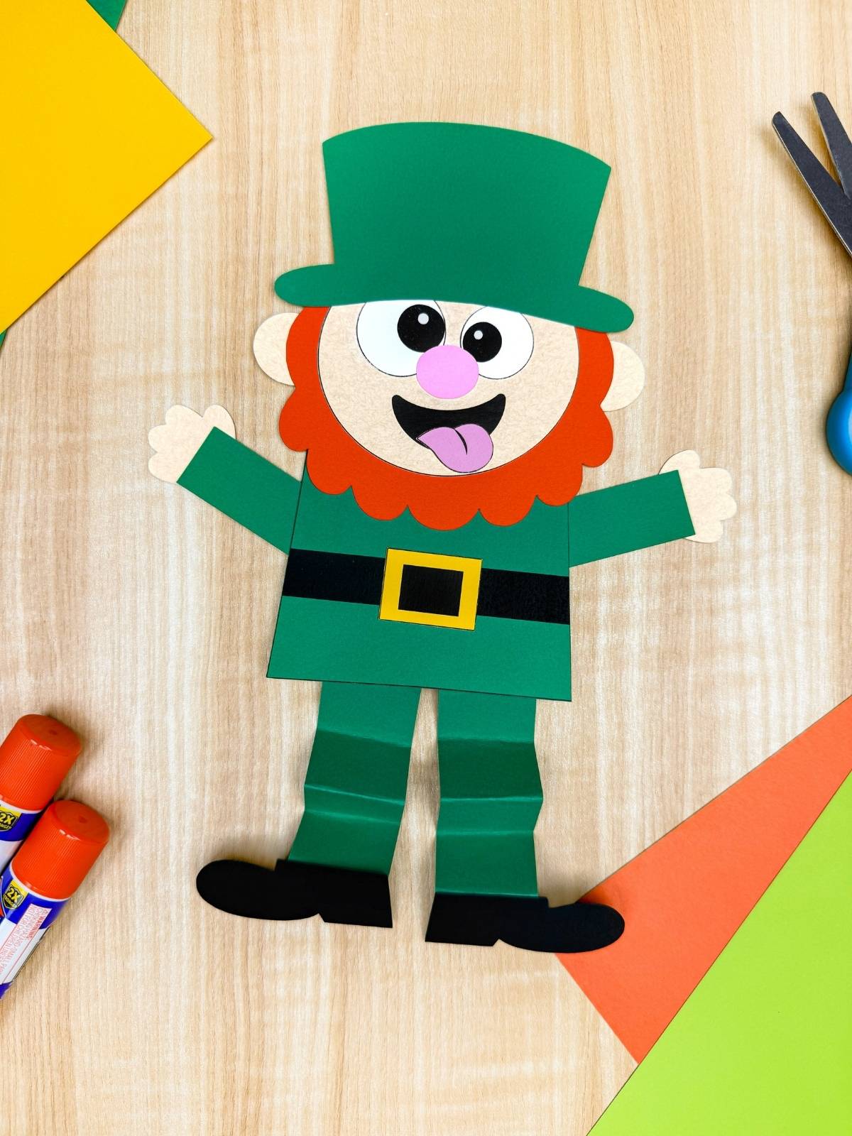A completed craft of a silly leprechaun paper craft with green hat, orange beard, and accordion legs on a craft table, fun and easy St. Patrick’s Day kids craft activity.