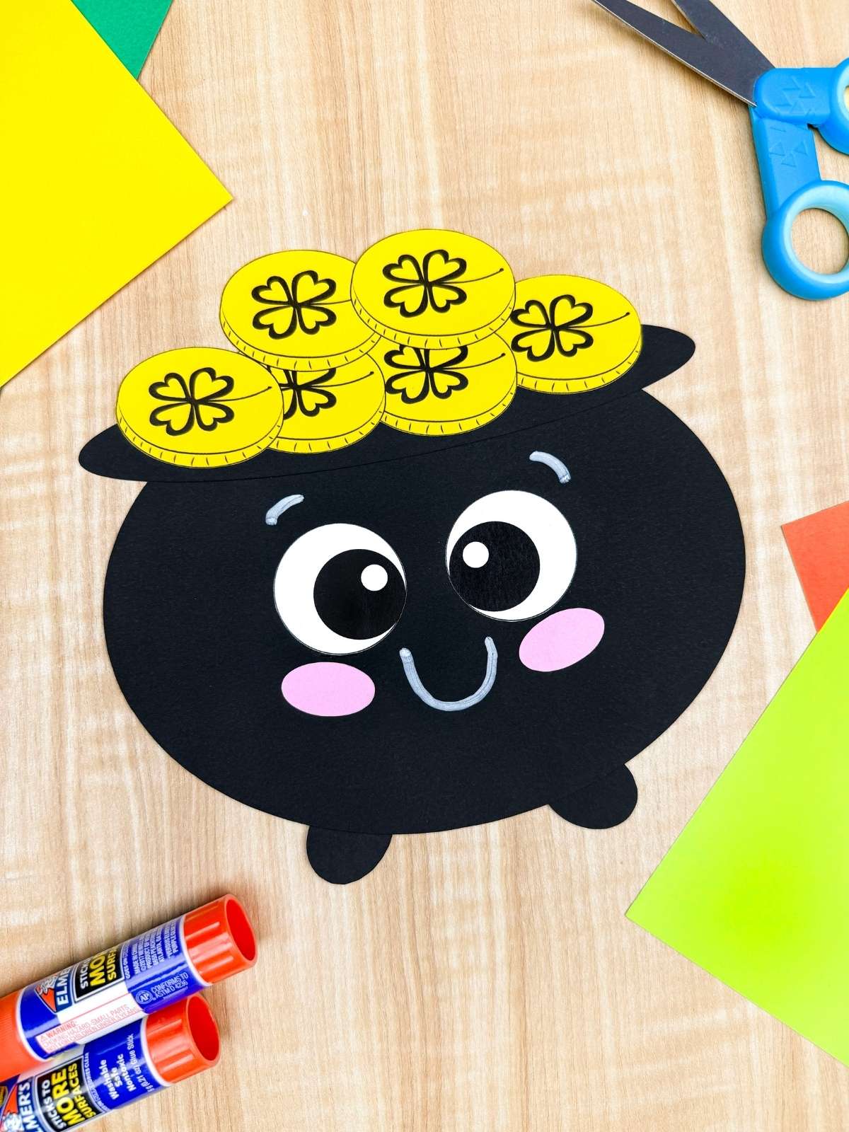 A completed craft of a smiling pot of gold shape craft with yellow coins and craft supplies for a St. Patrick’s Day kindergarten paper craft activity.