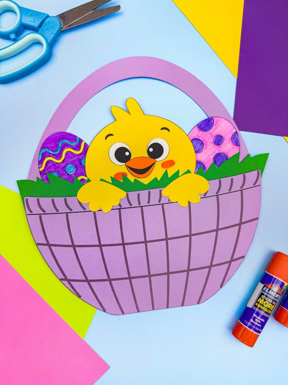 A completed craft chick Easter basket with colorful eggs surrounded by paper supplies for kids spring craft activity
