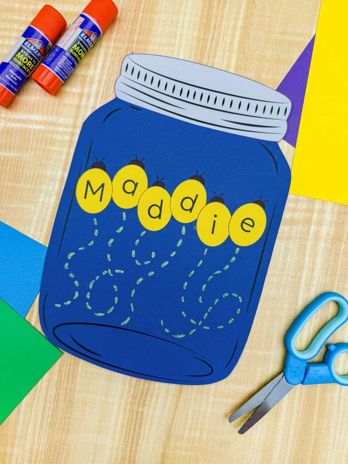A completed craft showing a blue mason jar firefly name craft with letters spelling Maddie surrounded by craft supplies