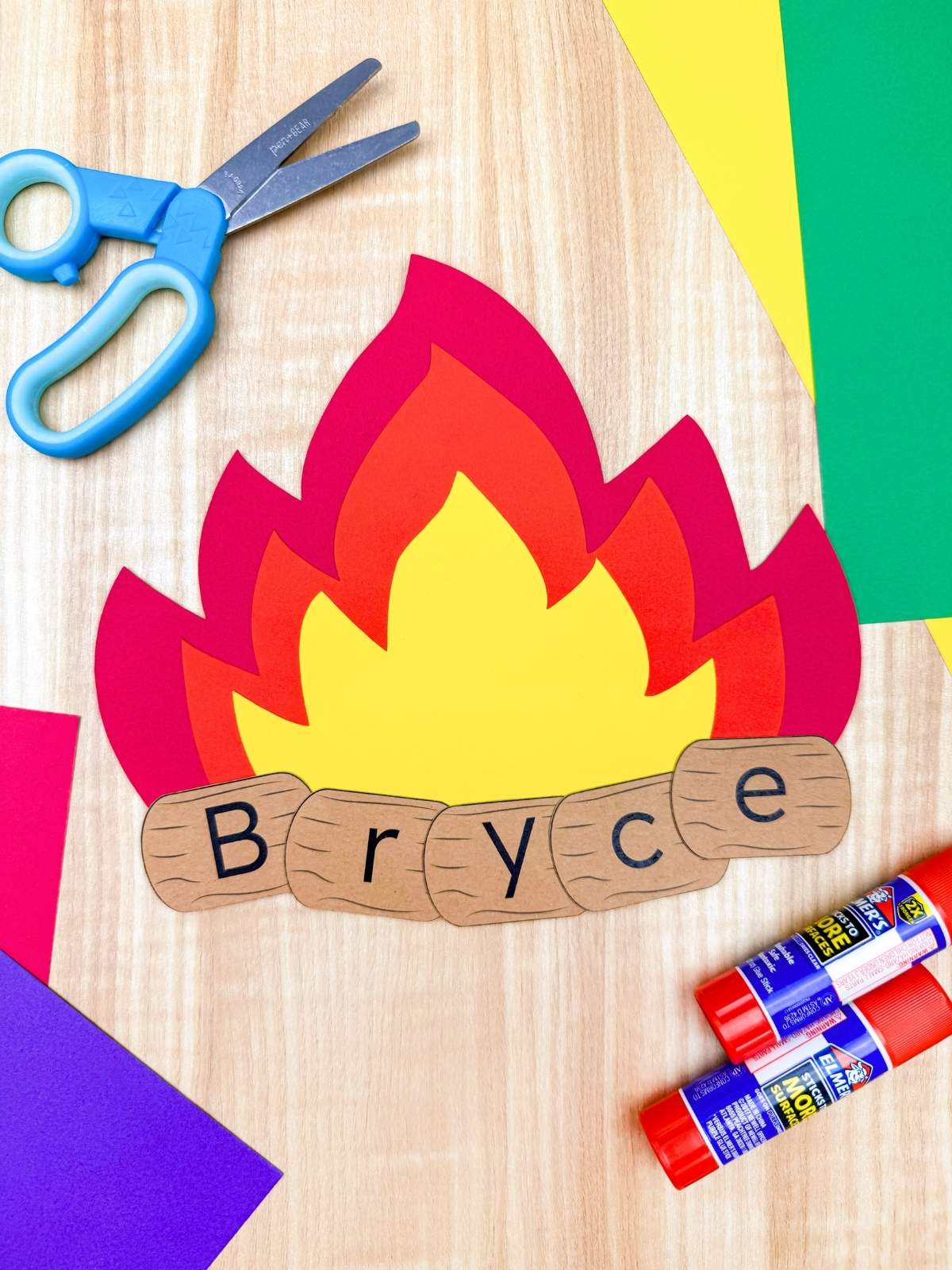A completed craft campfire name project with layered paper flames and the name Bryce spelled on log pieces with scissors and glue nearby