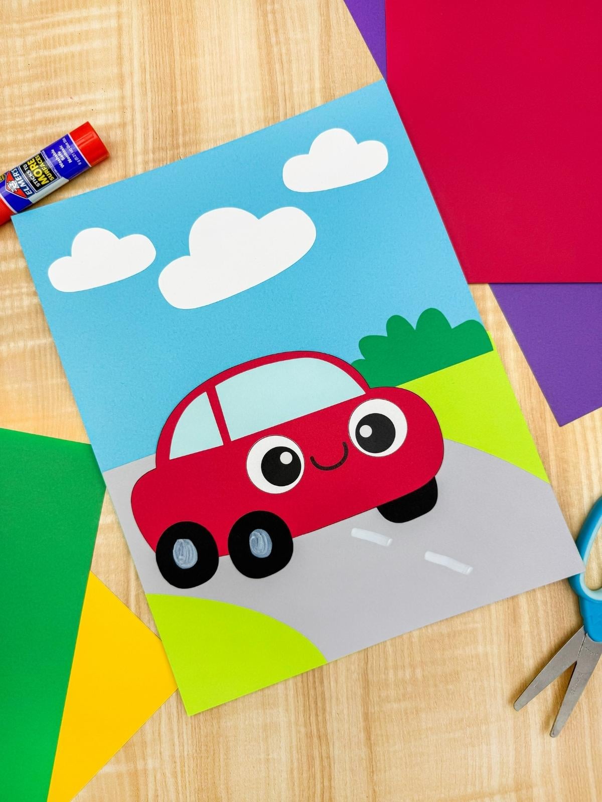 A completed craft of a red paper car on a road scene with clouds surrounded by colorful paper and scissors