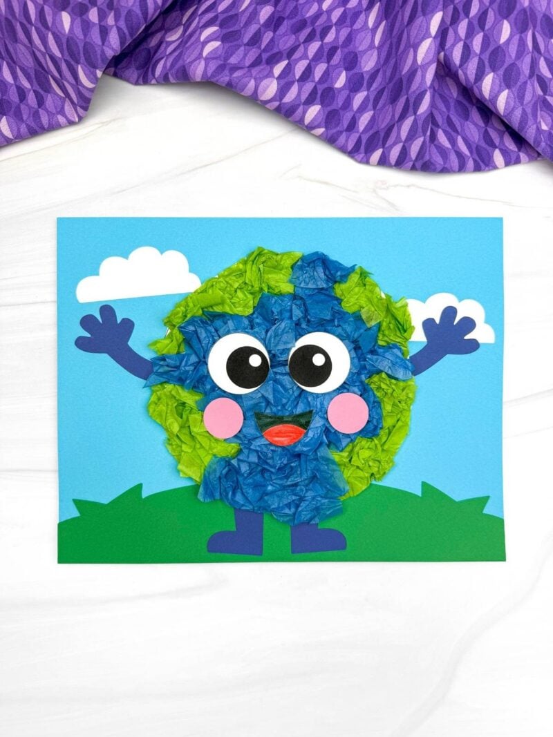 A completed craft of Earth tissue paper kids craft with smiling face on blue sky and green grass background for Earth Day activity