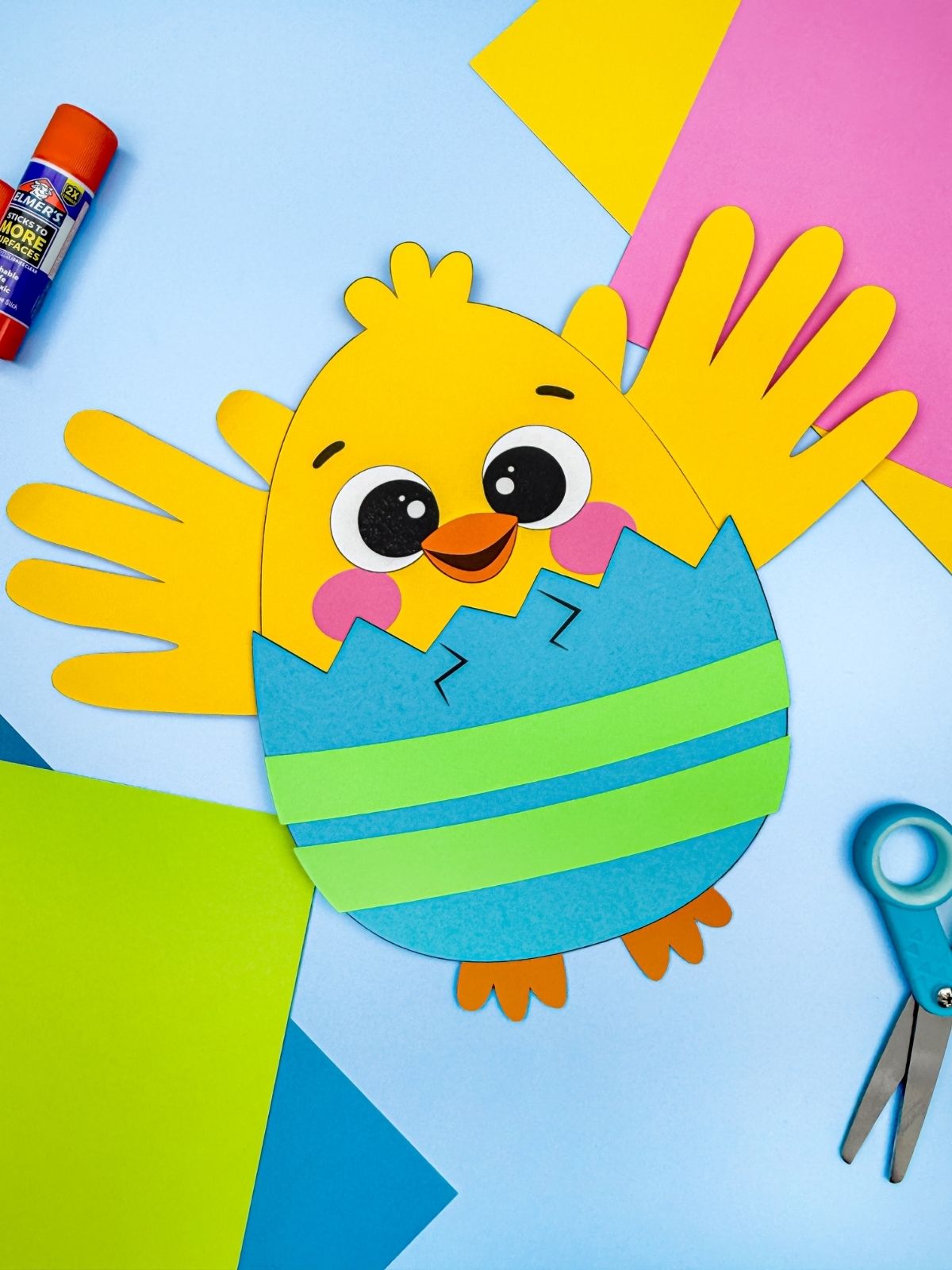 A completed Craft of a handprint Easter chick in a blue and green cracked eggshell with wings and feet surrounded by colorful paper scissors and glue for a kids Easter activity