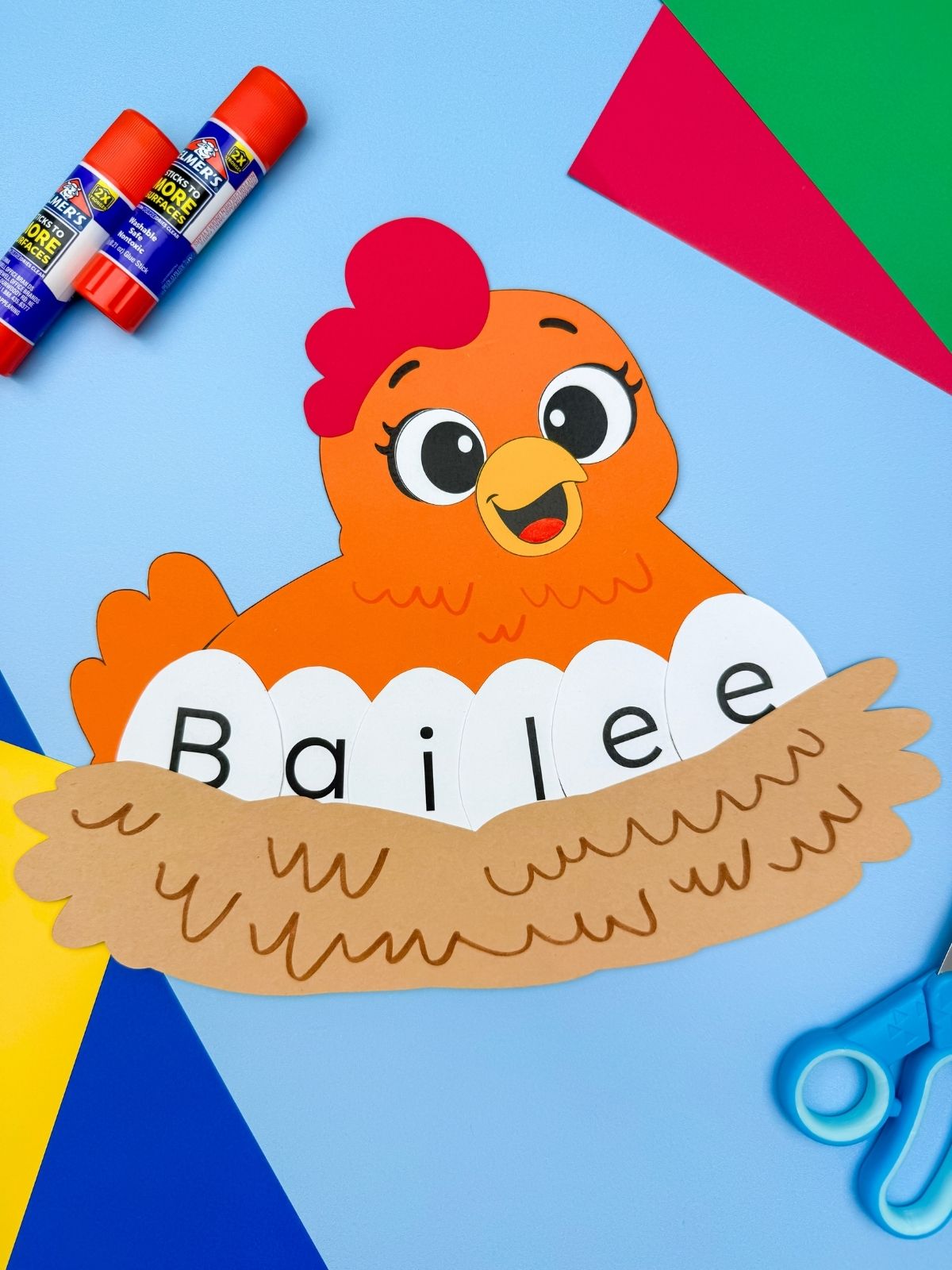 A completed chicken name craft with paper chicken, nest, and letter eggs spelling Bailee with scissors, glue, and colorful paper on a blue background