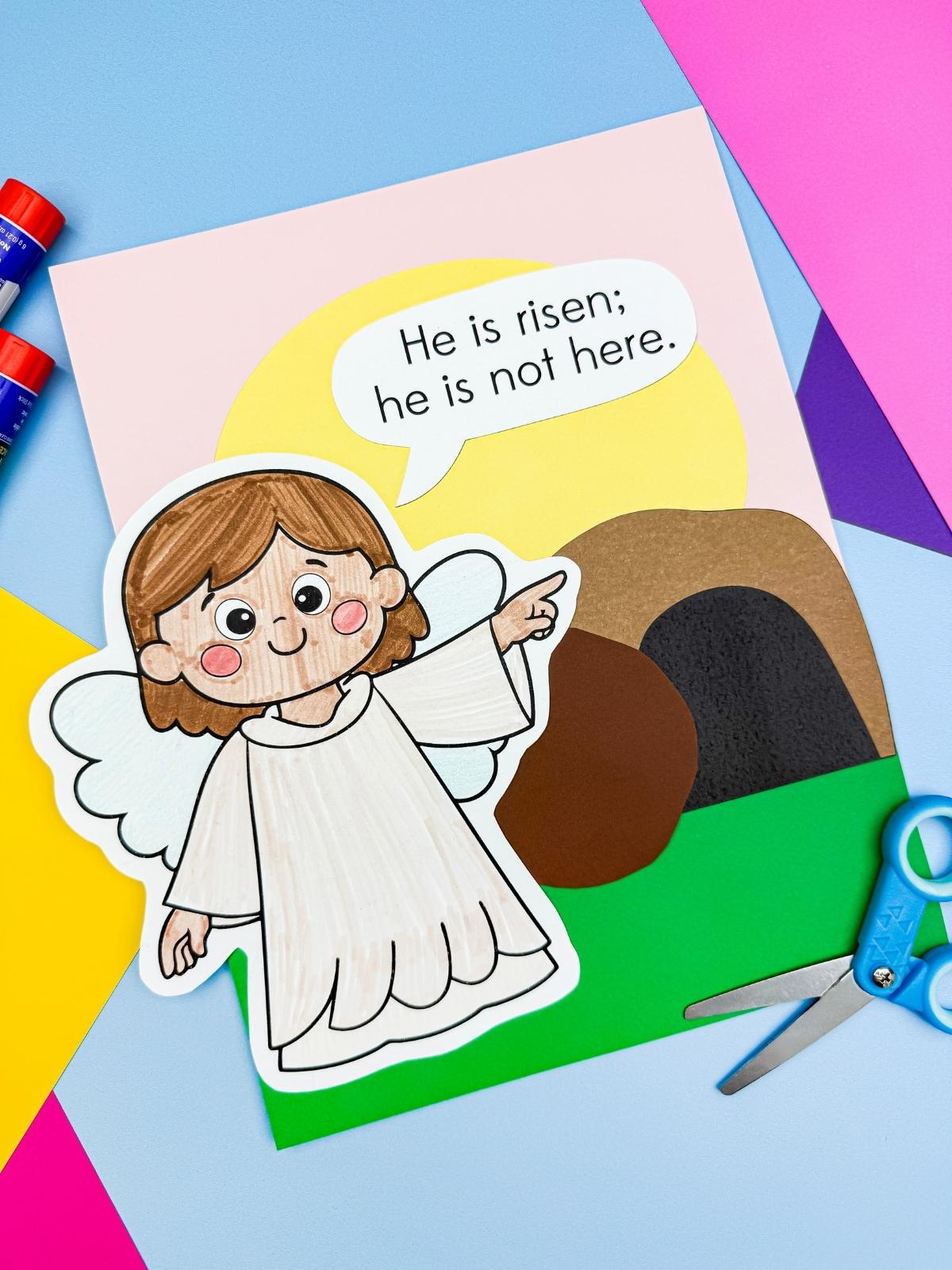 A completed craft showing an Easter resurrection Bible scene with angel pointing to the empty tomb and speech bubble saying He is risen he is not here.