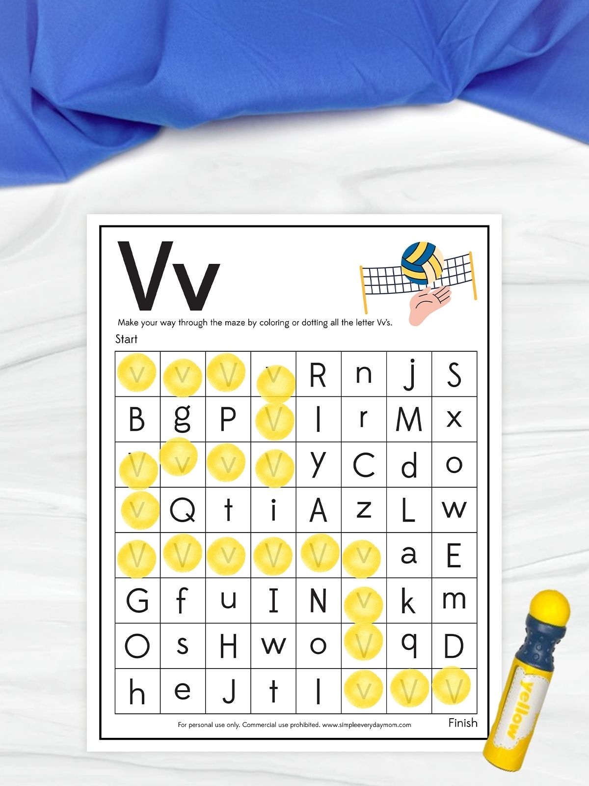 The image shows a children’s worksheet focused on the letter “V” with a maze-style activity. Kids are instructed to find and mark all the letter V’s to create a path from start to finish, and several of the correct letters are highlighted in yellow. A small volleyball illustration appears at the top, and a yellow marker is placed beside the page on a light surface with a blue fabric background.