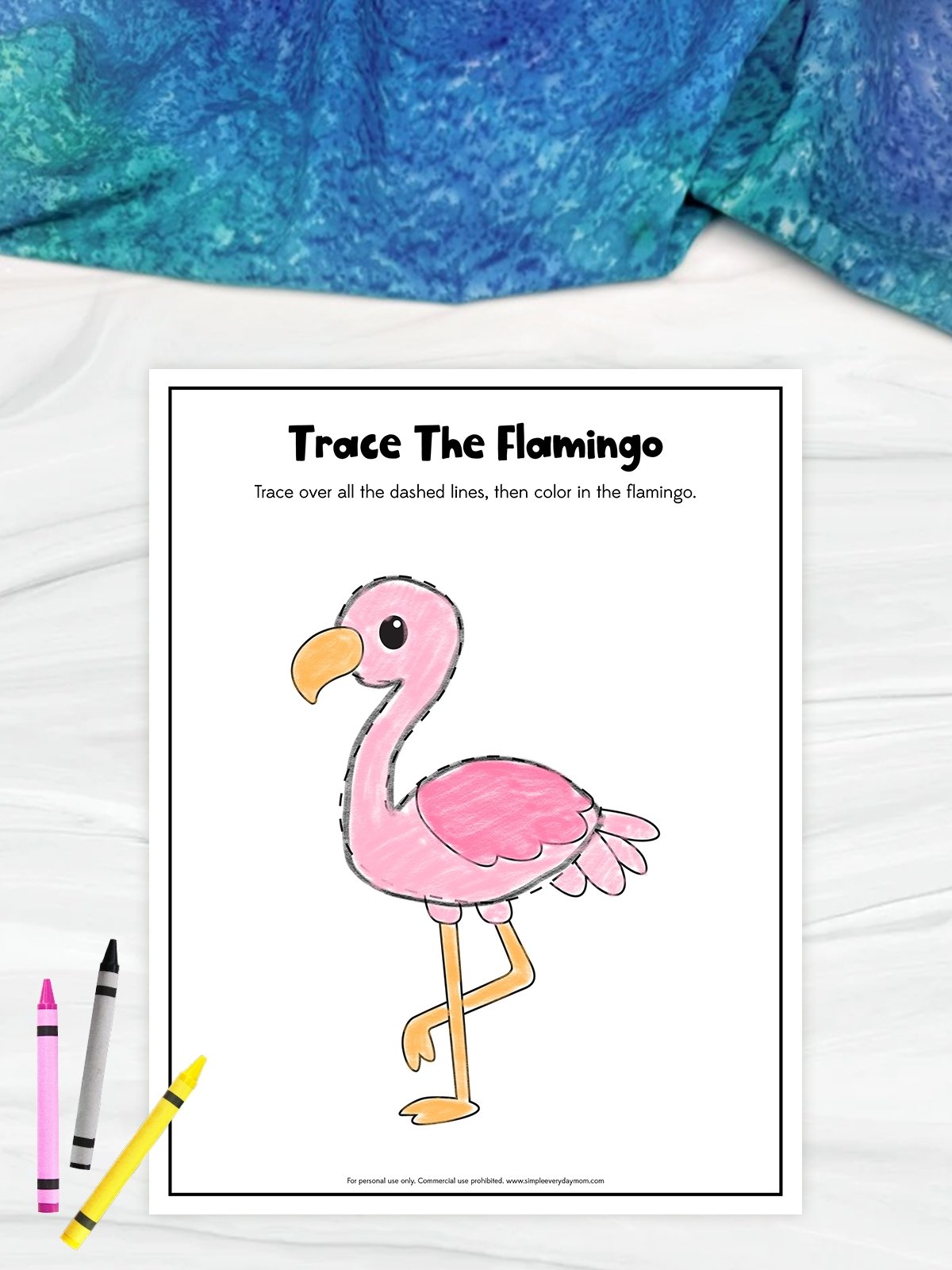 The image shows a children’s tracing worksheet titled “Trace The Flamingo” on a light surface with a blue fabric background. It features a large flamingo drawing outlined with dashed lines for tracing, which has been partially colored in pink and orange. A few crayons are placed beside the page, emphasizing the coloring activity.