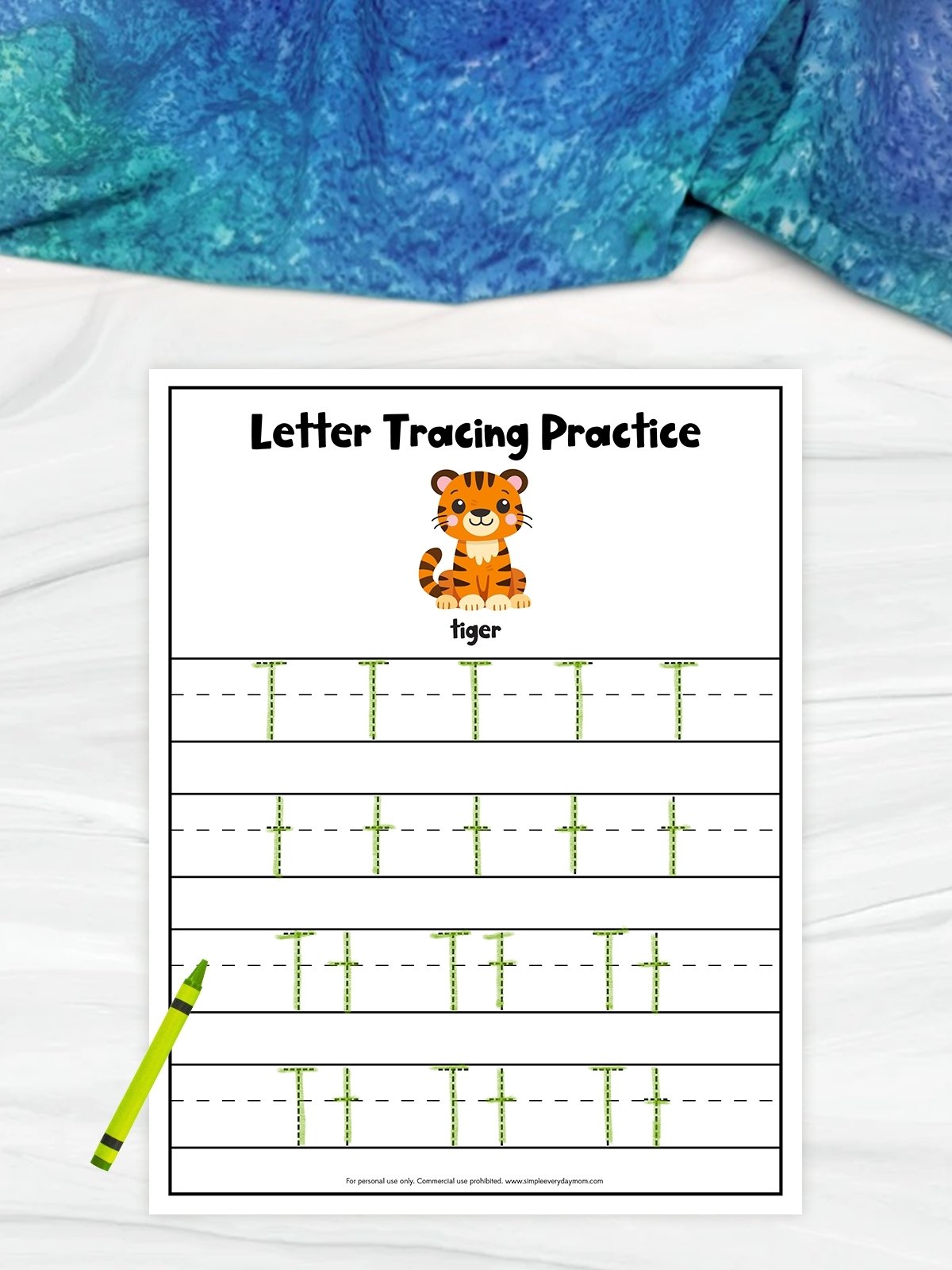 The image shows a children’s letter tracing worksheet focused on the letter “T.” At the top is a cute cartoon tiger with the word “tiger,” followed by rows of dashed uppercase and lowercase T’s for tracing practice. A green crayon lies beside the page, and the background includes a light surface with blue fabric above.