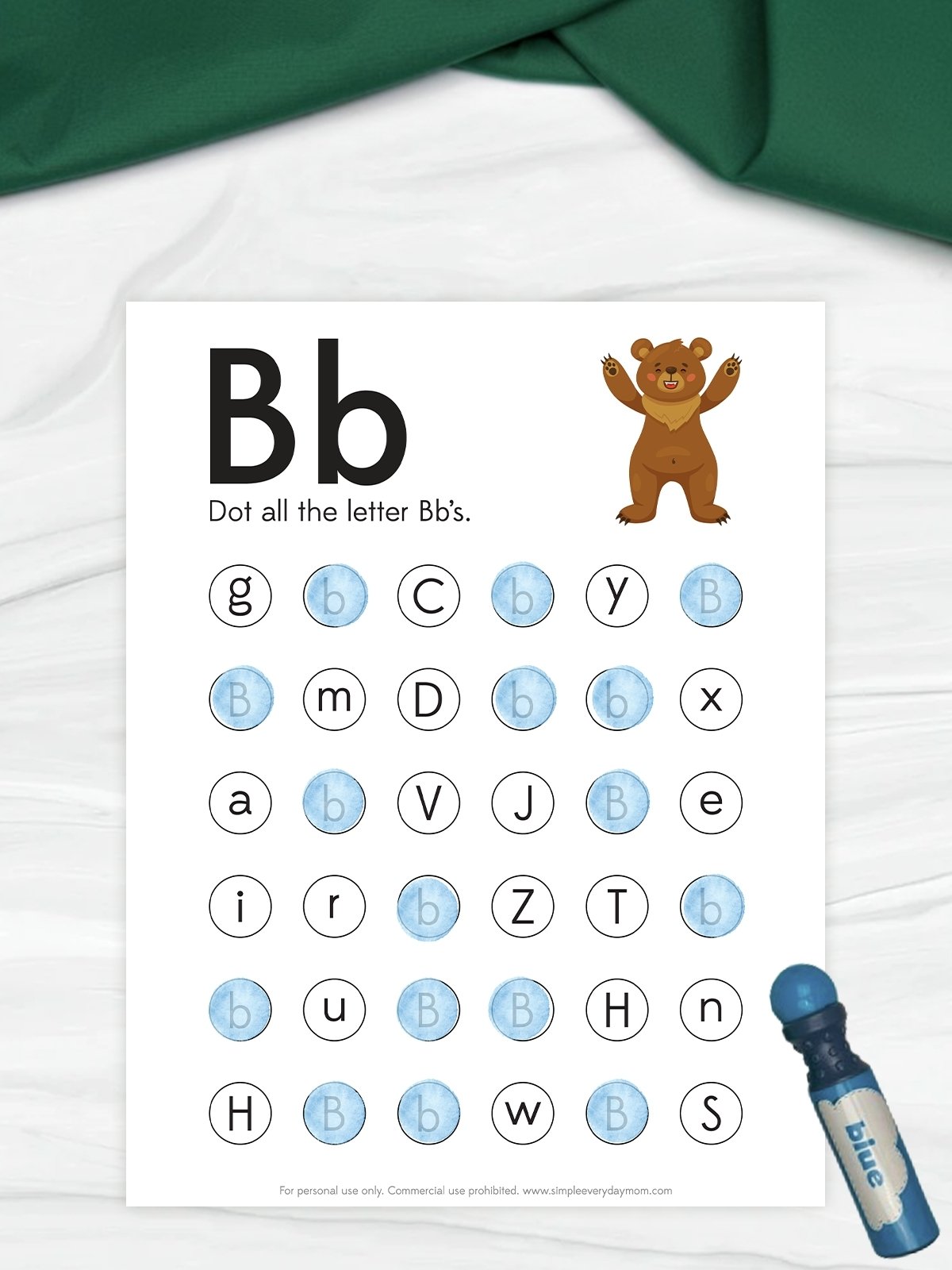 The image shows a children’s worksheet focused on the letter “Bb,” with instructions to dot all the letter B’s. A grid of mixed letters is displayed, with several B’s already marked in blue using a dot marker. A small bear illustration and a blue dot marker beside the page add a fun, hands-on learning element.
