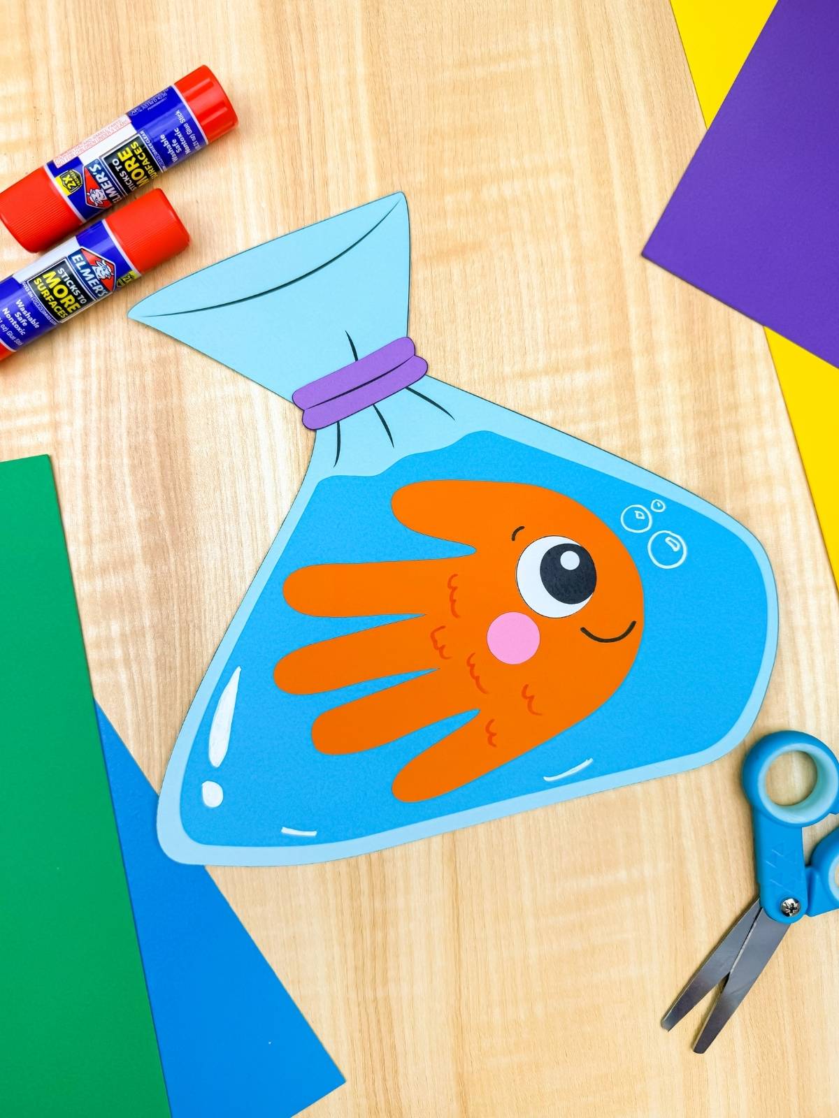 A completed craft showing a handprint goldfish inside a blue paper bag design with glue sticks, scissors, and colorful paper on a wooden surface
