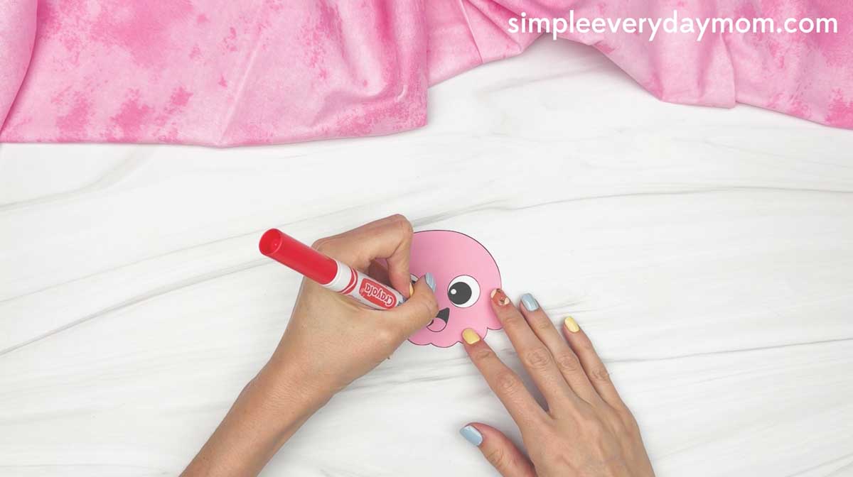 Hands coloring a pink octopus cutout with a red marker on a white table.