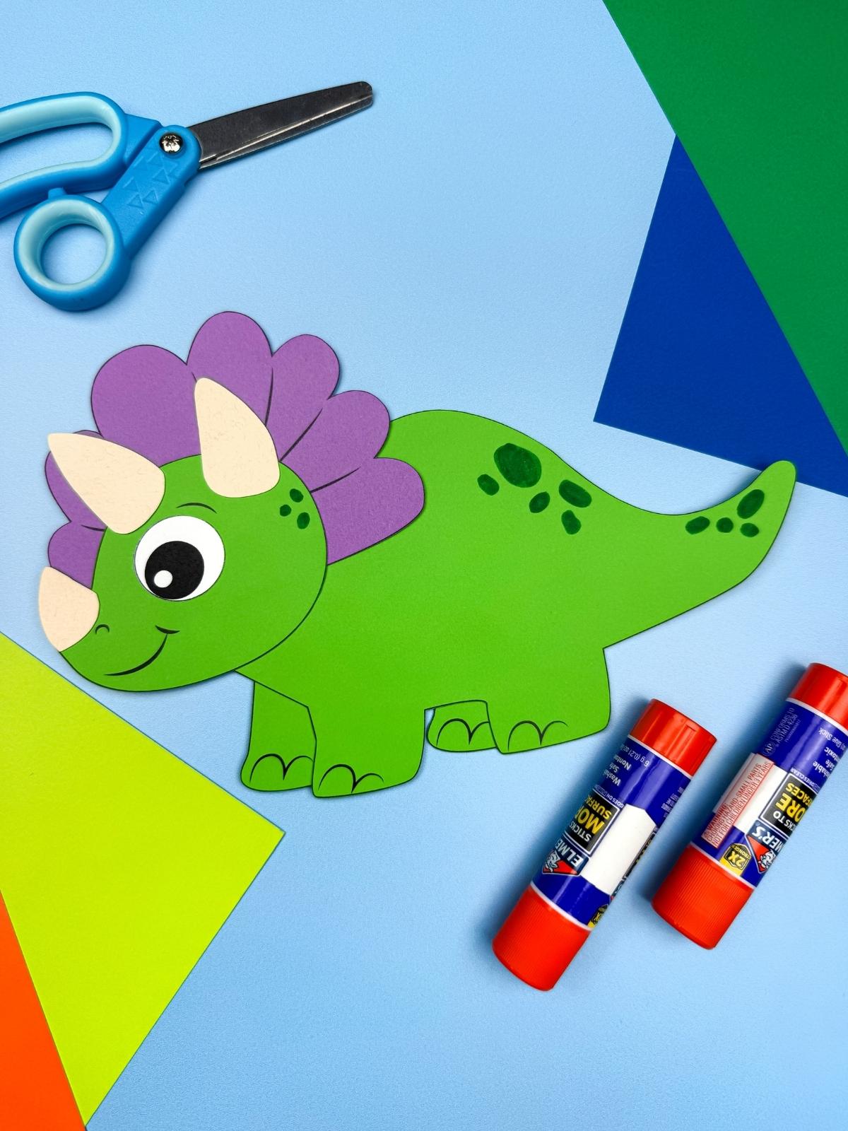 A completed craft showing a green triceratops dinosaur paper craft with purple frill surrounded by scissors, glue sticks, and colorful paper on a blue background