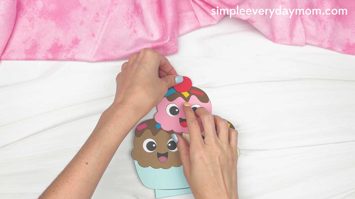Hands placing a red cherry on top of a smiling ice cream sundae craft.