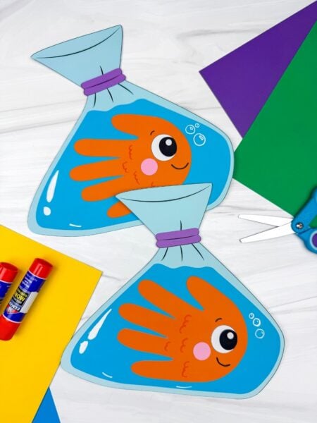 A completed craft showing two handprint goldfish in blue paper bag designs with colorful paper, glue sticks, and scissors nearby