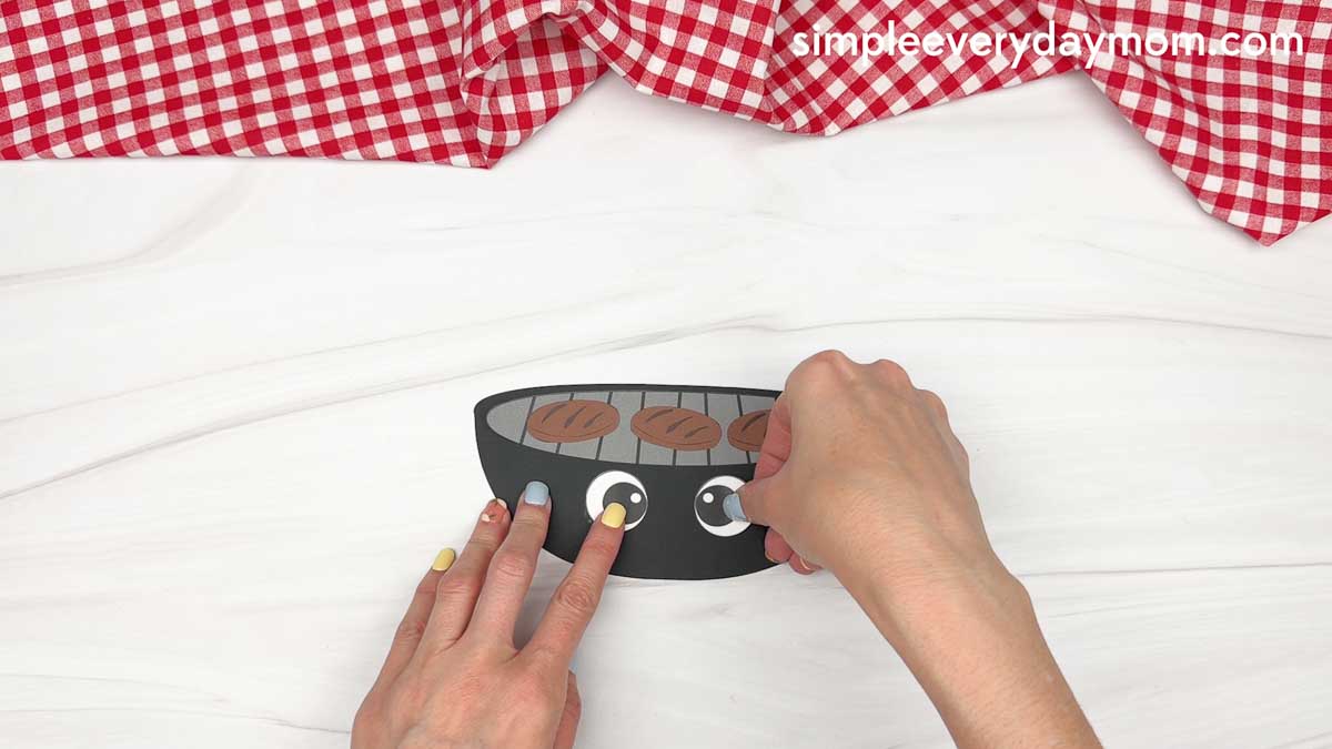 Hands attaching cartoon eyes to the grill base with burger patties for a Father’s Day craft
