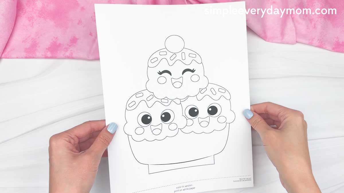 Hands holding a printable ice cream sundae coloring page with three smiling scoops.