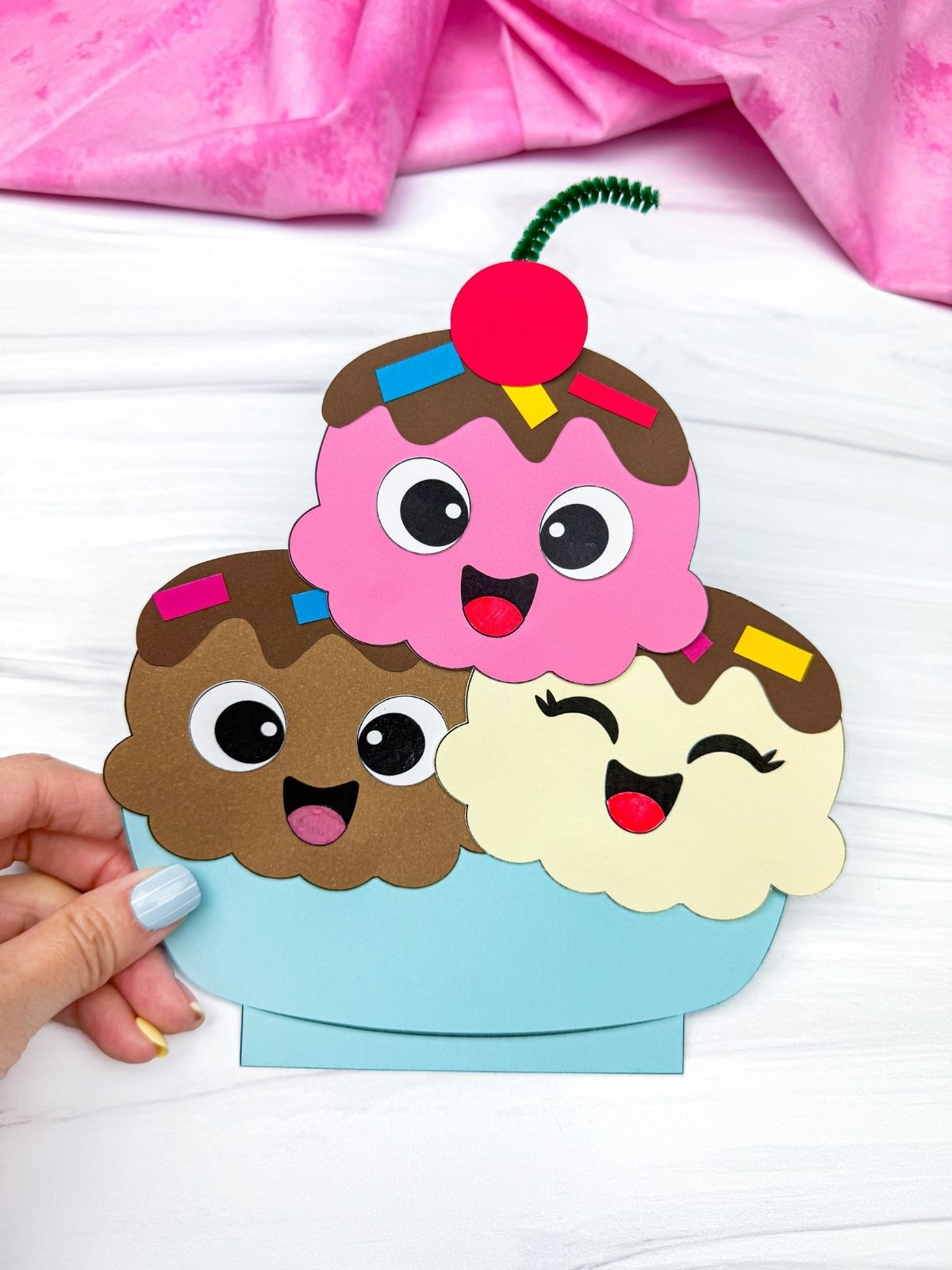 Hands holding a completed ice cream sundae craft with three smiling scoops and a cherry on top