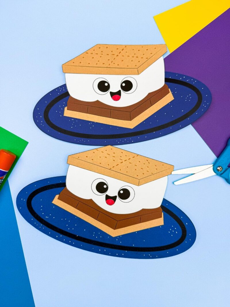 A completed craft of two smiling s’mores on blue bases with craft supplies nearby