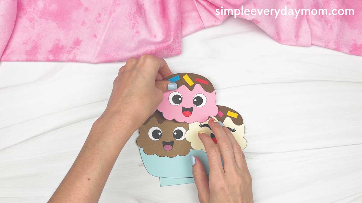 Hands assembling colorful ice cream scoops with sprinkles onto a sundae base.