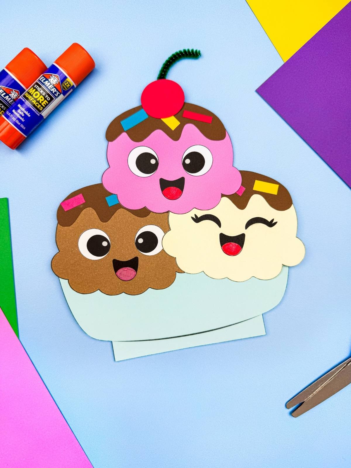A completed craft of a smiling ice cream sundae with three scoops surrounded by craft supplies