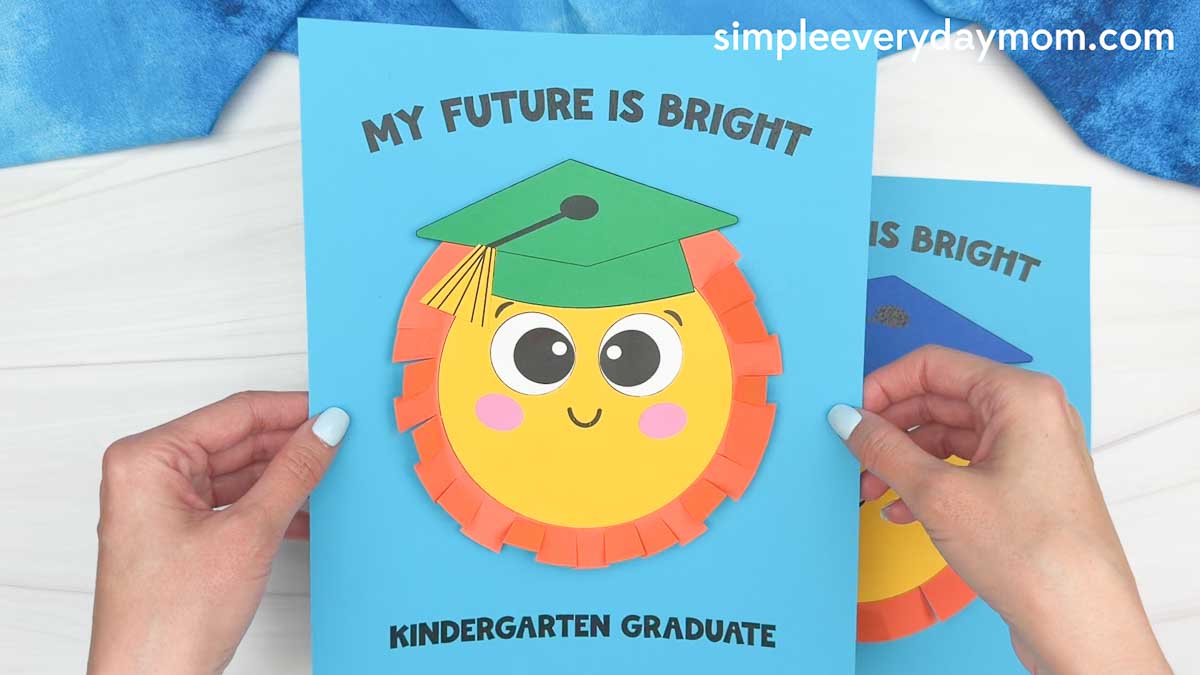 Hands holding a completed sun graduation craft card with a smiling sun wearing a graduation cap and the words “My Future Is Bright” and “Kindergarten Graduate” on blue paper