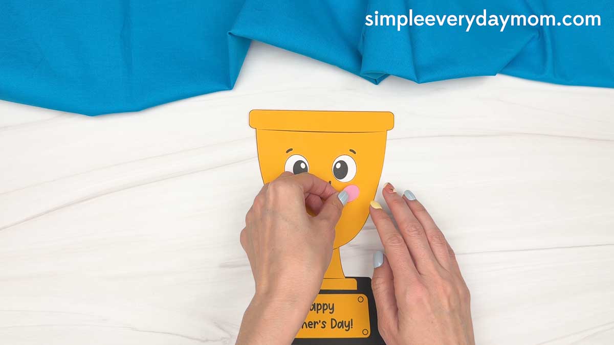 Hands placing the pink cheek pieces onto the smiling Father’s Day trophy craft.
