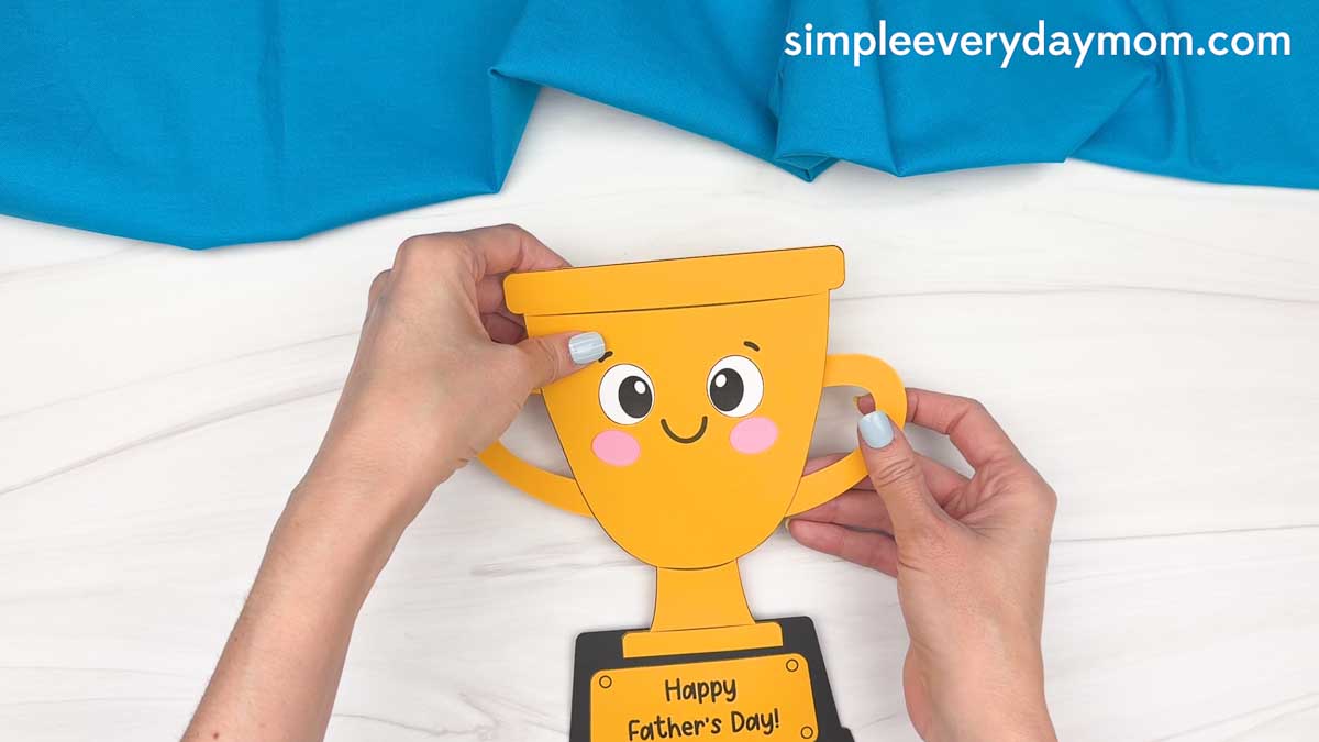 Hands attaching the handles to the completed smiling Father’s Day trophy craft.