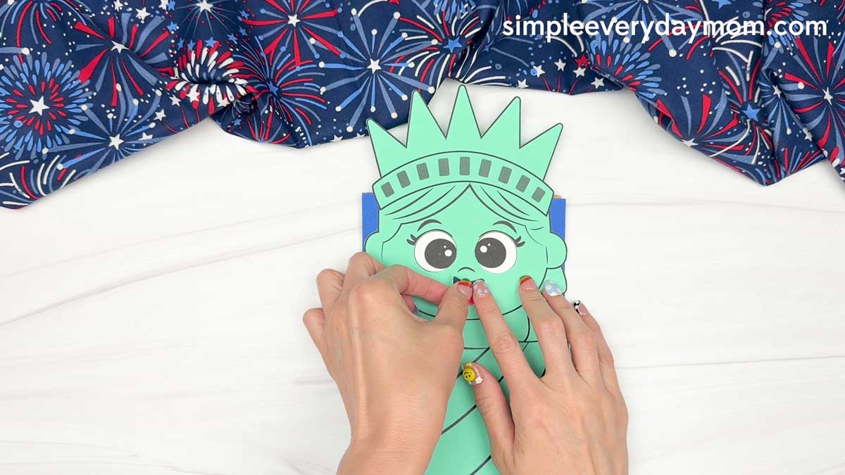 Hands attaching the nose piece to the Statue of Liberty paper bag puppet craft face.