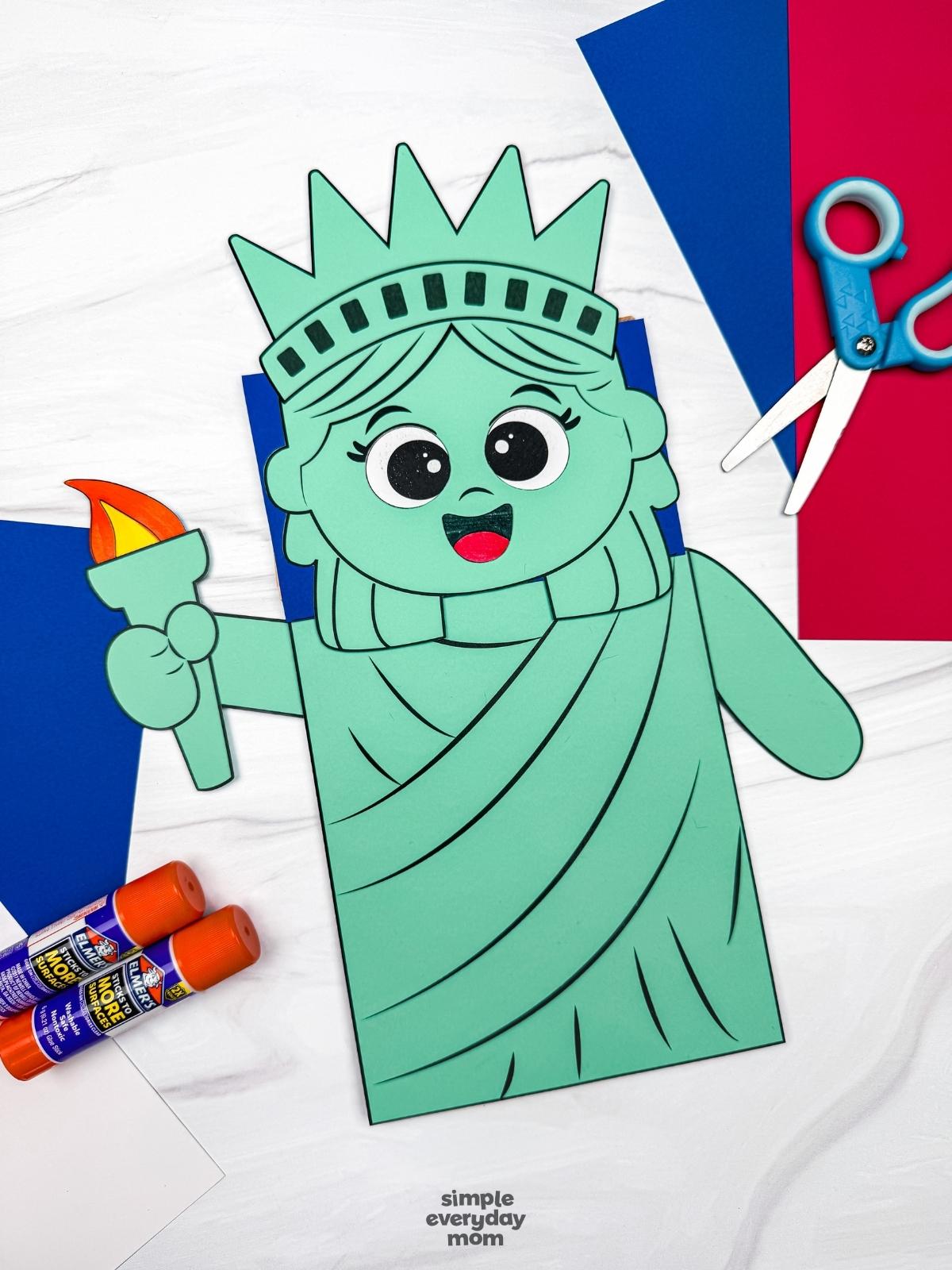A completed craft showing a Statue of Liberty paper bag puppet surrounded by colored paper, scissors, and glue sticks.