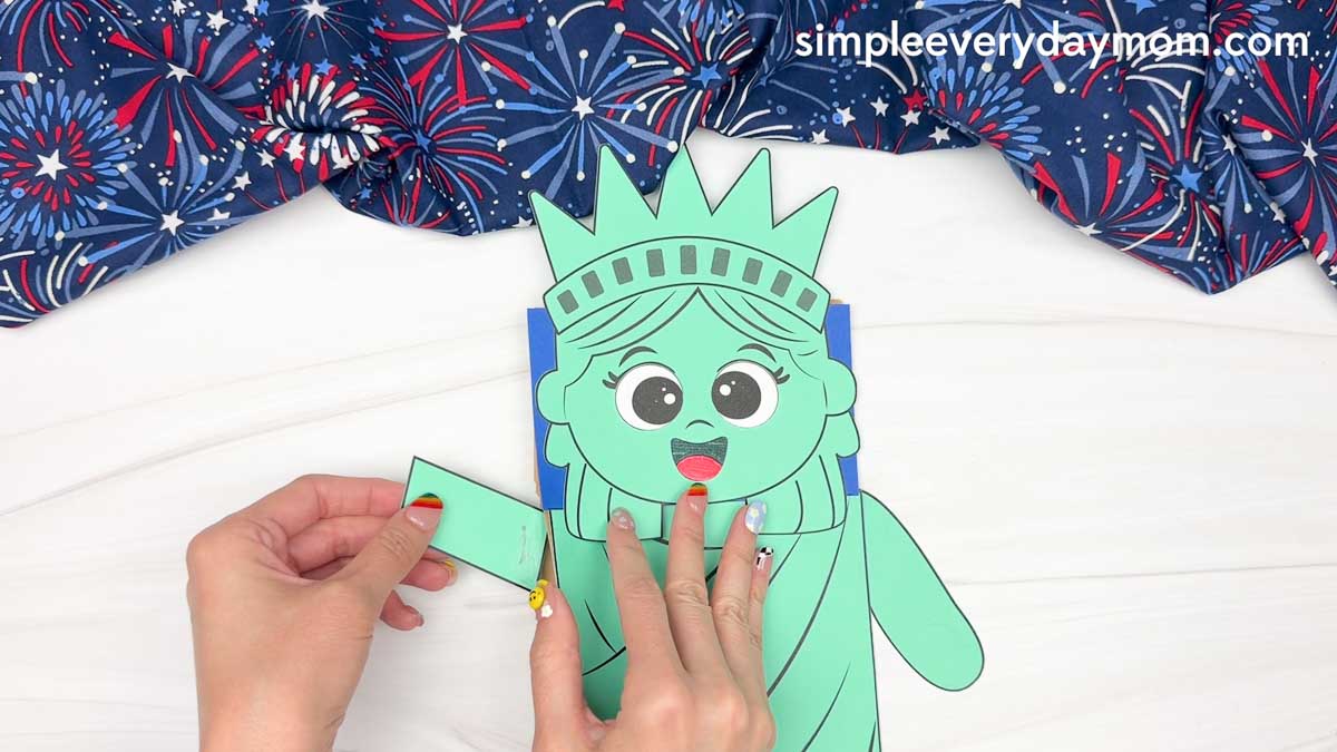 Hands attaching an arm piece to the Statue of Liberty paper bag puppet craft with the face and body already assembled.