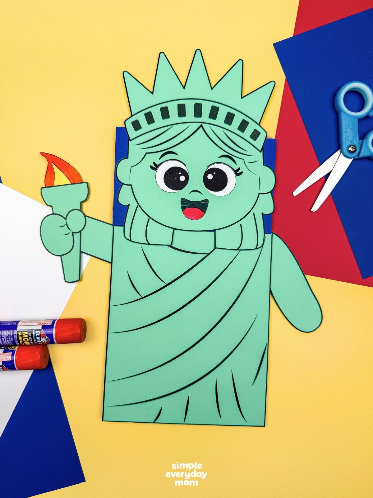 A completed craft showing a Statue of Liberty paper bag puppet with craft supplies on a yellow background.