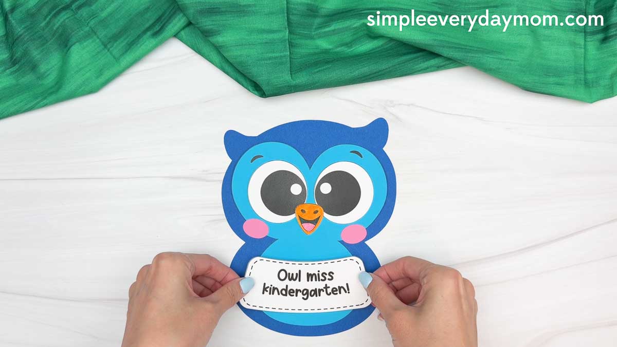 Hands placing the “Owl miss kindergarten!” sign onto the owl graduation craft.