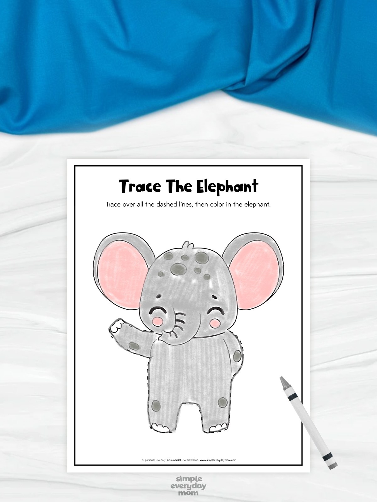This image features a printable “Trace The Elephant” worksheet created for preschool children to practice tracing and coloring skills. The worksheet shows a cute cartoon elephant with dashed outline lines that children can trace before coloring the picture. A gray crayon beside the page and the soft blue fabric background give the activity a calm, playful, and child-friendly appearance.