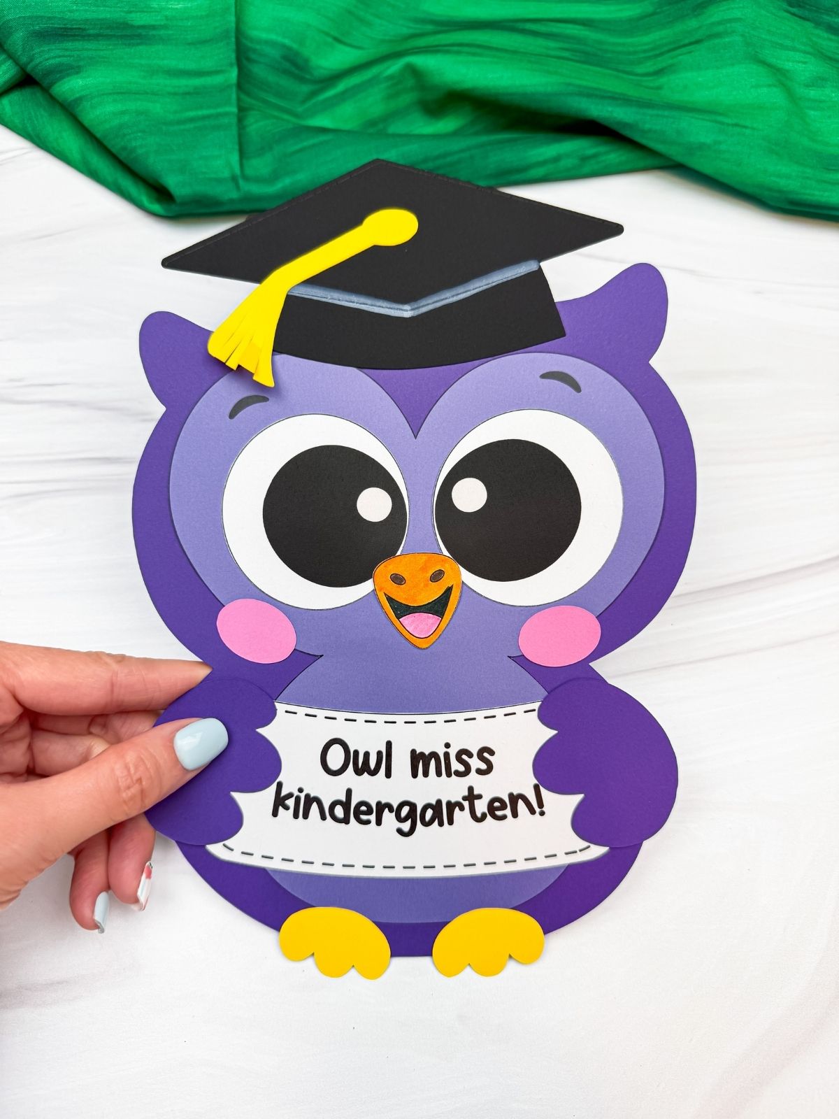 Hands holding a completed purple owl graduation craft with a graduation cap and “Owl miss kindergarten!” sign.