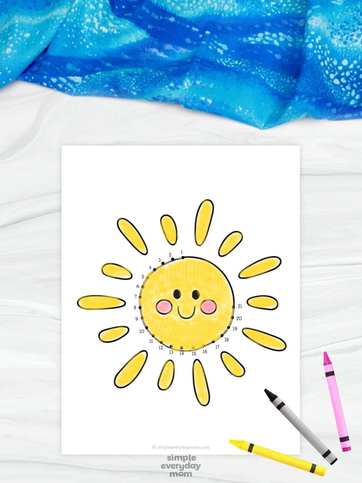 The image shows a beach-themed connect-the-dots worksheet featuring a smiling yellow sun with rosy cheeks and bright sun rays. The numbered dots around the sun help children trace and complete the picture as part of a fun educational activity. Colored crayons and a soft blue fabric background add a playful and creative classroom feel to the design.