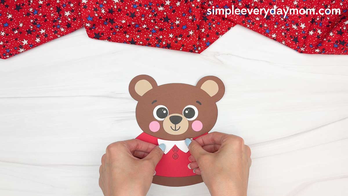 Hands attaching a white collar piece to a brown teddy bear paper craft for a patriotic teddy bear activity