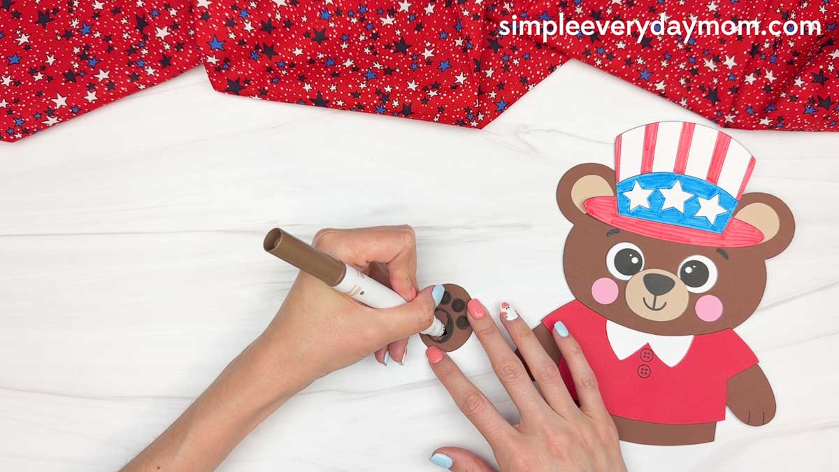 Hands coloring the teddy bear paw piece with a brown marker beside a completed patriotic teddy bear craft activity