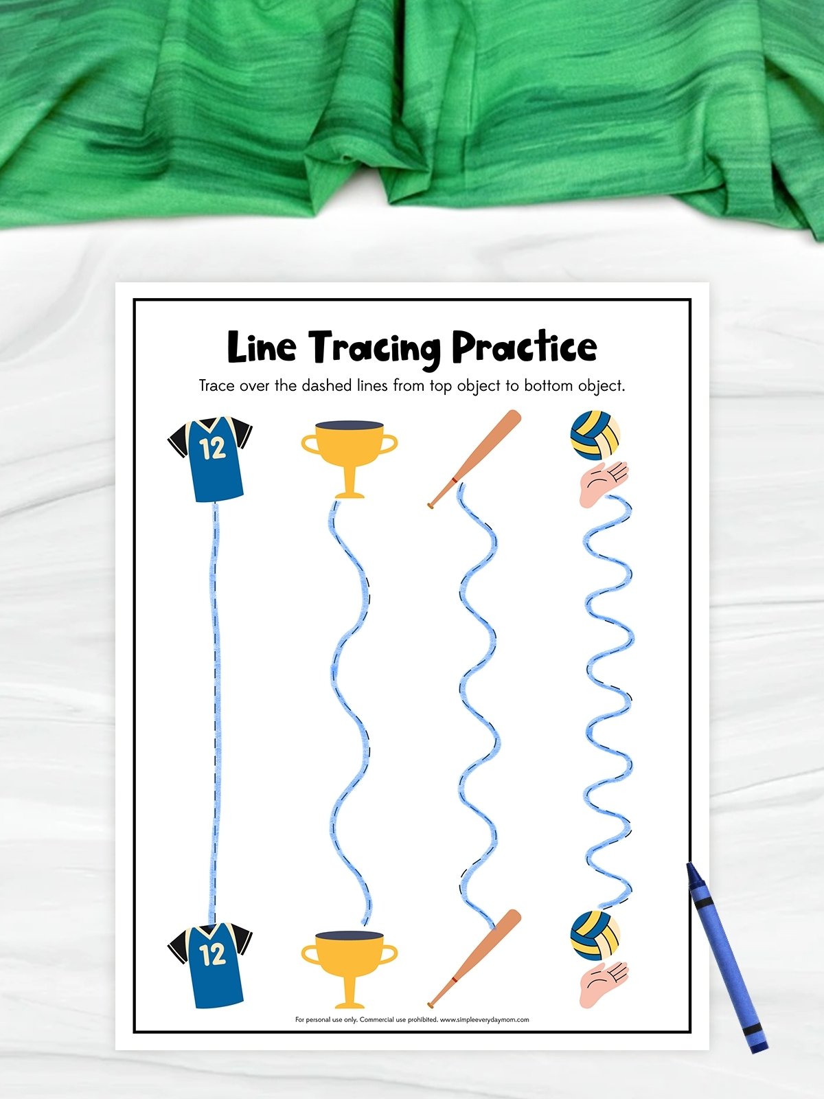 The image shows a children’s worksheet titled “Line Tracing Practice” with a sports theme. Kids are encouraged to trace different dashed lines connecting sports-related objects like a jersey, trophy, baseball bat, and volleyball from top to bottom. A blue crayon is placed beside the worksheet, and a green cloth is visible in the background.