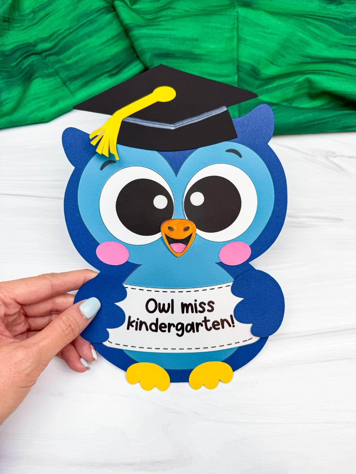 Hands holding a completed blue owl graduation craft with a graduation cap and “Owl miss kindergarten!” sign.