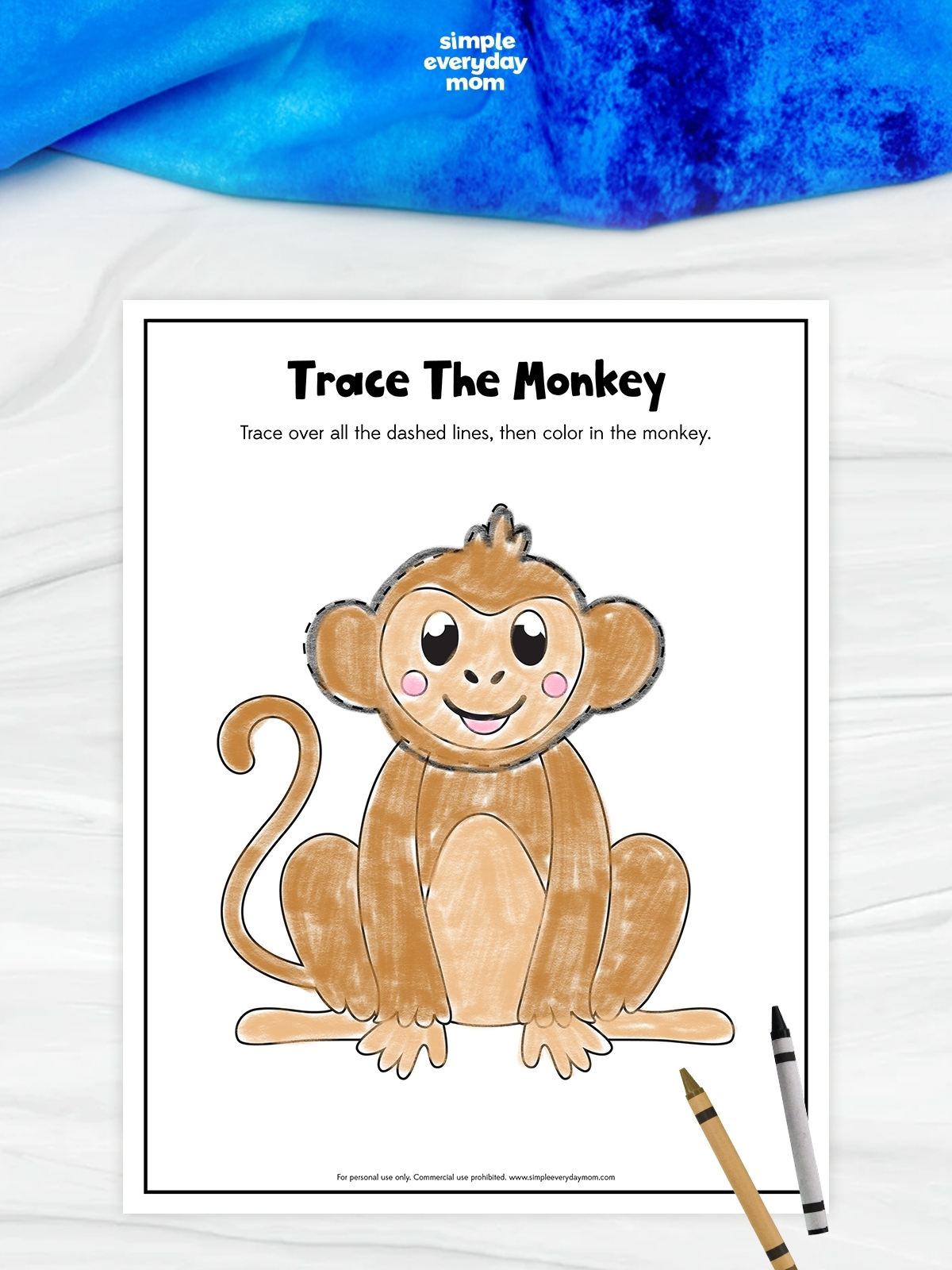 This image features a preschool worksheet titled “Trace The Monkey.” The activity asks children to trace over the dashed outline of a smiling cartoon monkey and then color it in. The monkey is colored light brown with pink cheeks and sits with its curled tail to the side. Two crayons are placed near the bottom corner of the page. The worksheet is designed to help young children practice tracing, hand control, and coloring skills.