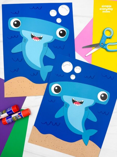 A completed hammerhead shark craft displayed on blue underwater backgrounds alongside colorful craft supplies, scissors, and glue sticks.