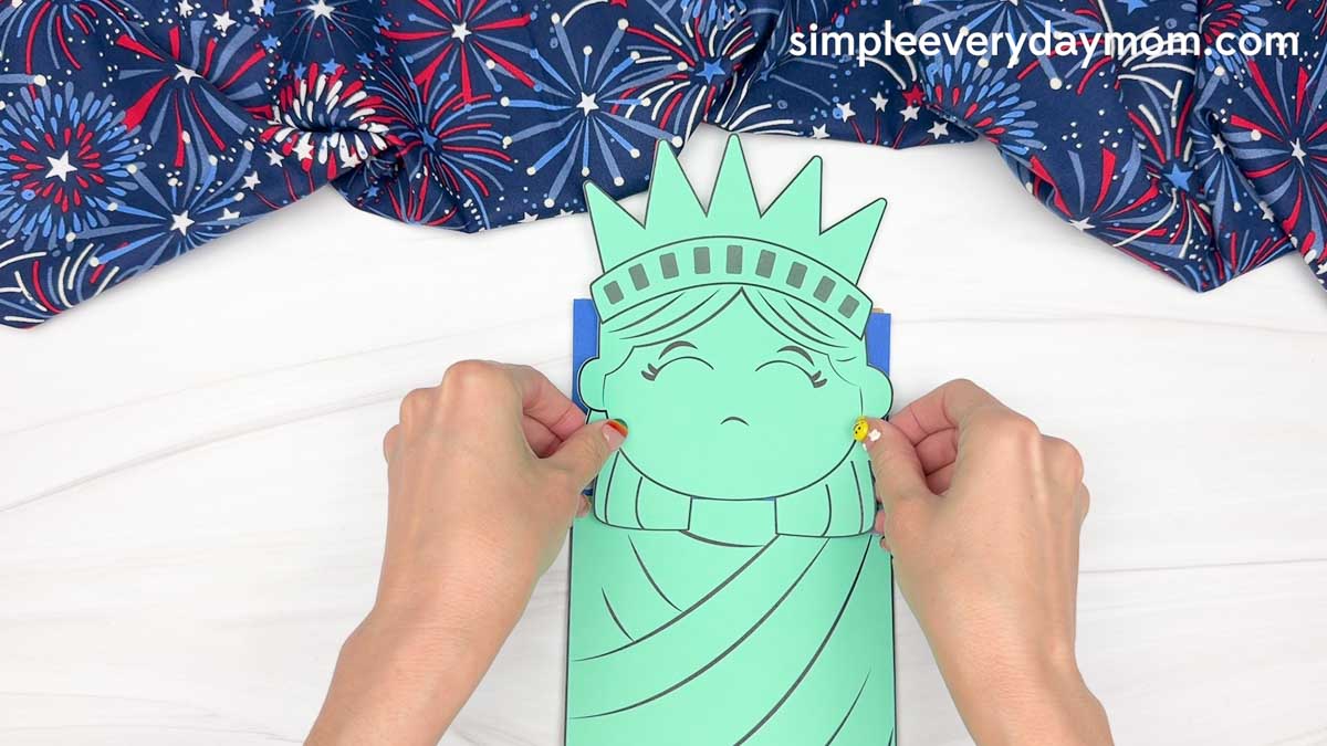 Hands placing the Statue of Liberty head cutout onto the paper bag puppet craft above the body piece.